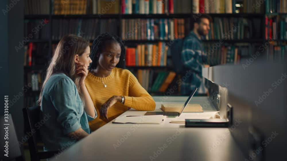University Library: Two Gifted Girl Students Study, Use Laptop Computer, Help Each Other with Advice, Working on Assignment and Preparing for Exams. Focused Smart Student Learning, Studying for Exams University Library: Two Gifted Girl Students Study, Use Laptop Computer, Help Each Other with Advice, Working on Assignment and Preparing for Exams. Focused Smart Student Learning, Studying for Exams