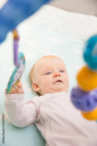 Cute adorable baby girl playing with hanging rattle. Six month little child lying in bed. Childhood or baby care concept