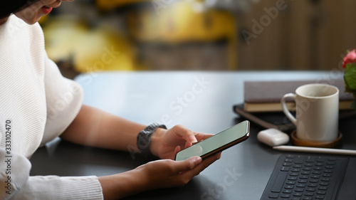 Cropped image of young creative woman s hands holding a smartphone with empty screen while sitting at the working desk over comfortable living room as background.