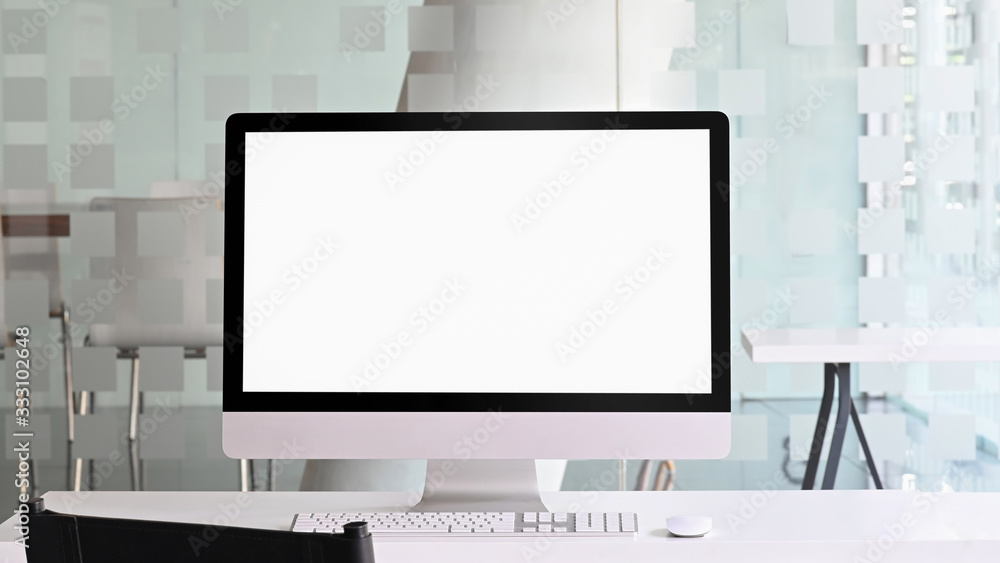 Fototapeta premium Photo of computer monitor with white blank screen putting on white working desk with wireless mouse and keyboard over orderly office as background.