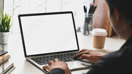 Cropped image of businessman hands typing on computer laptop with white blank screen that putting on orderly working desk that surrounded with books, pencil holder, coffee cup and potted plant.