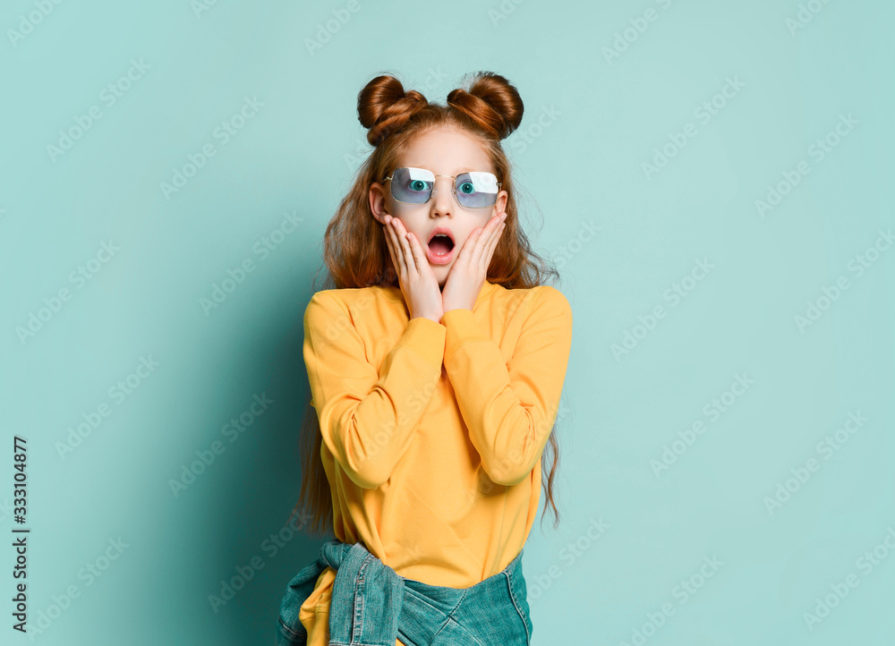 Shocked scared red-haired teen girl in stylish smoked glasses and yellow sweatshirt gasps covering her cheeks with hands
