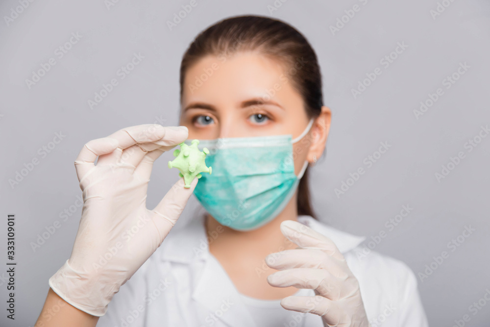 Close-up model of coronavirus in the hands of a doctor. A young woman in a white coat, mask and gloves studies the virus.