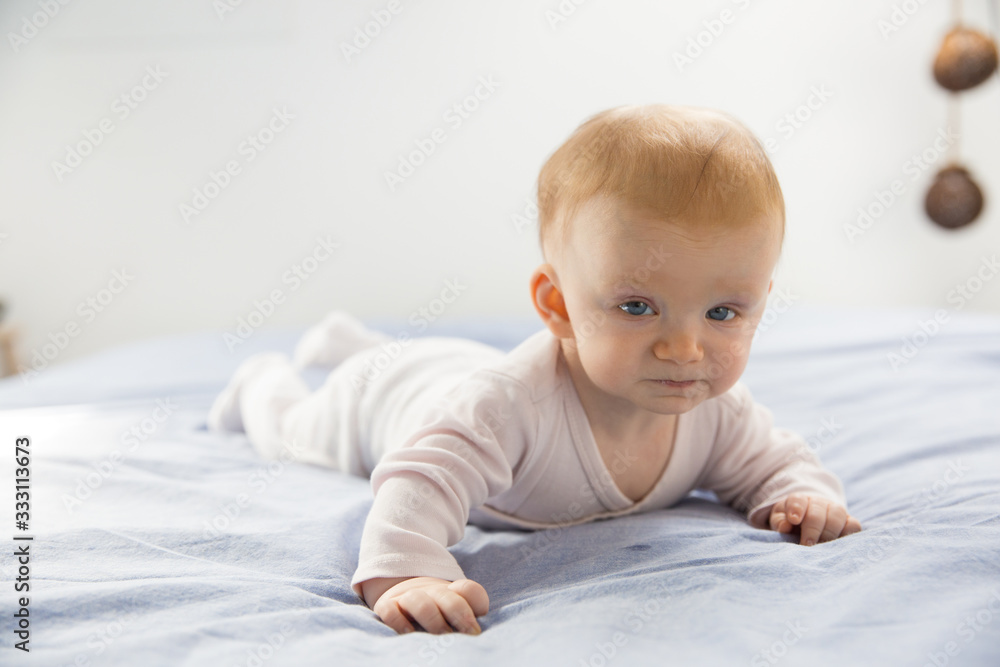 Serious funny adorable baby girl crawling in bedding. Six month child lying on belly at home. Childhood or baby care concept