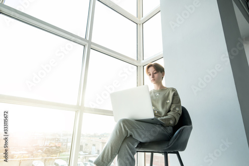 A teenage girl is sitting in a chair with a laptop near the panoramic window