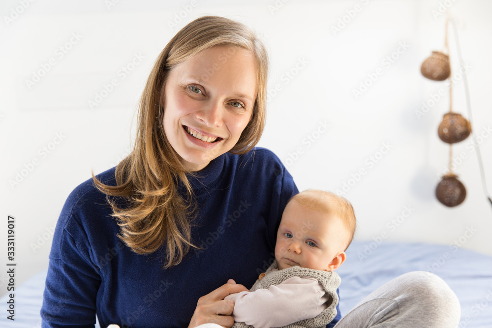 Happy cheerful new mother holding cute baby girl in arms, looking at camera, smiling. Young woman nursing six month daughter in bedroom at home. Motherhood concept Happy cheerful new mother holding cute baby girl in arms, looking at camera, smiling. Young woman nursing six month daughter in bedroom at home. Motherhood concept