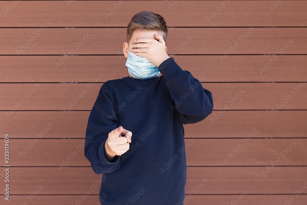 A boy standing outside with protection face mask shields eyes with his hand and points his finger