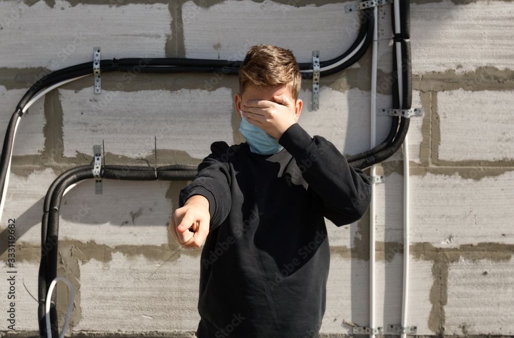 A boy standing outside with protection face mask shields eyes with his hand and points his finger