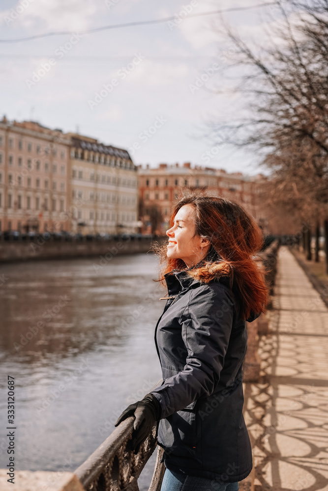 A brunette girl in a winter jacket stands on the embankment near the bridge with a view of the embankment, the Neva river and buildings