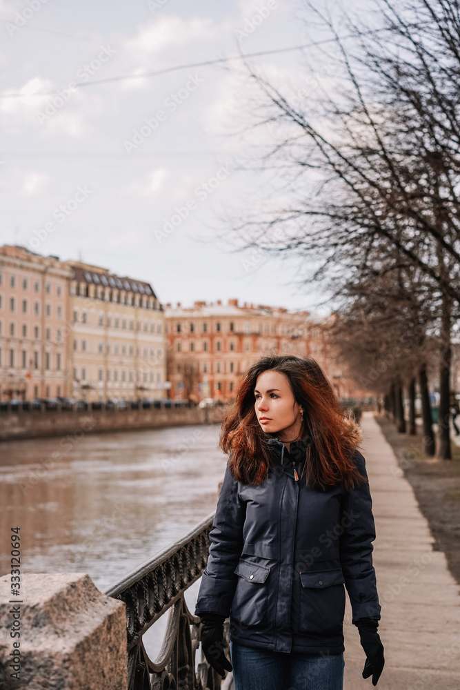 A brunette girl in a winter jacket stands on the embankment near the bridge with a view of the embankment, the Neva river and buildings