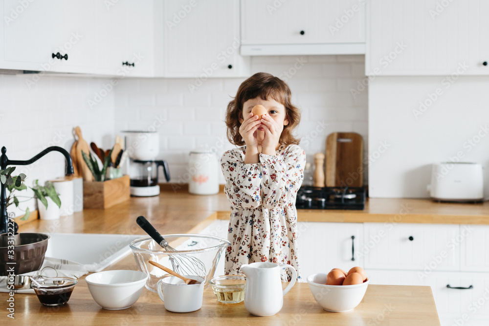 Cute 5 years girl wearing dress preparing cake at the white modern kitchen. Cute 5 years girl wearing dress preparing cake at the white modern kitchen.
