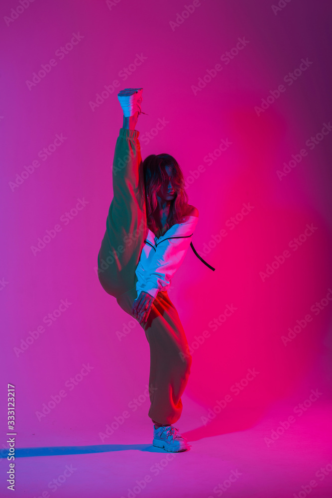 Fototapeta Fashion model young modern woman in stylish clothes in trendy gym shoes dancing indoors with bright multi-colored color. Sporty girl dancer posing in a room with bright neon glamour pink-blue light.