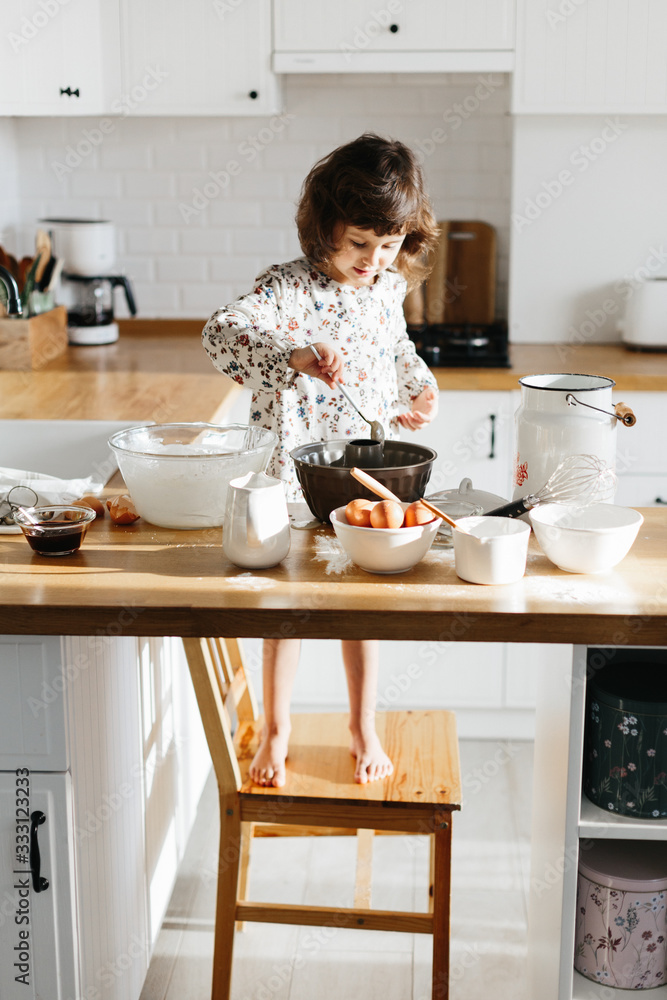 Cute 5 years girl wearing dress preparing cake at the white modern kitchen.