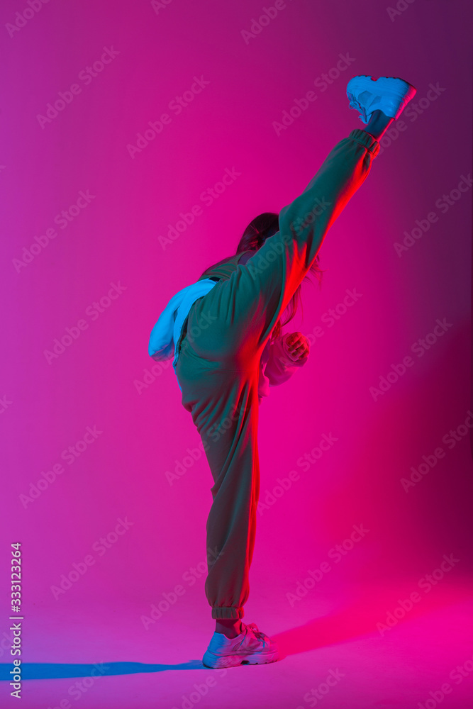 Fototapeta Pretty fashionable model young woman in stylish clothes in trendy gym shoes dancing indoors with bright colorful light. Sporty girl kicks in a studio with bright neon beauty pink-blue color