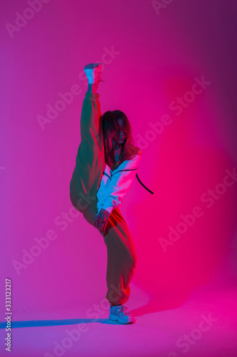 Fototapeta Fashion model young modern woman in stylish clothes in trendy gym shoes dancing indoors with bright multi-colored color. Sporty girl dancer posing in a room with bright neon glamour pink-blue light.