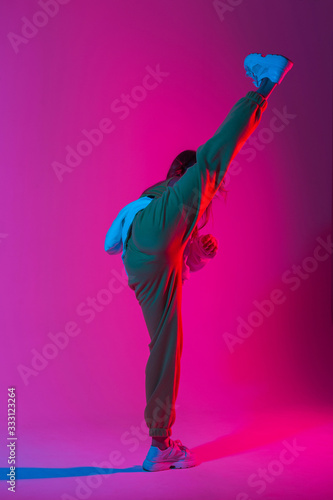 Fototapeta Pretty fashionable model young woman in stylish clothes in trendy gym shoes dancing indoors with bright colorful light. Sporty girl kicks in a studio with bright neon beauty pink-blue color