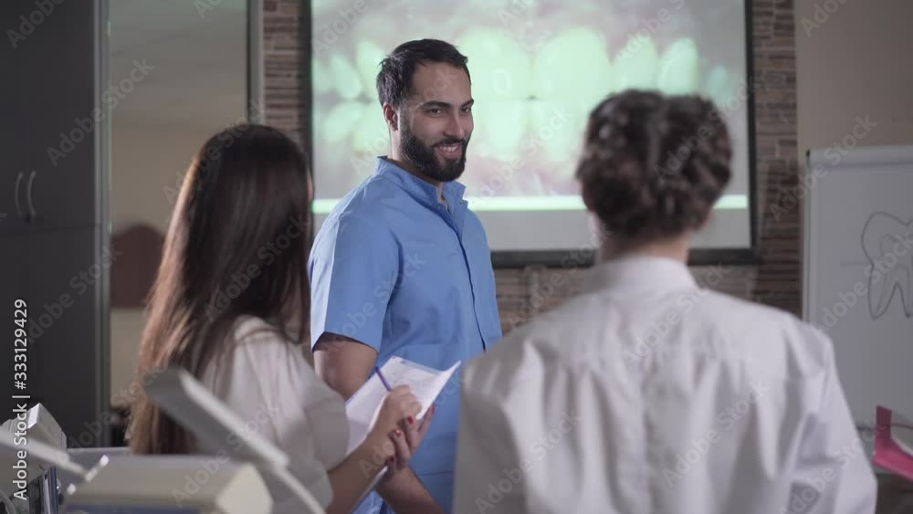 Smiling Middle Eastern lecturer talking in front of screen with teeth visualization. Confident young man giving lecture on dentistry to students. Medicine, teaching, odontology, stomatology. Smiling Middle Eastern lecturer talking in front of screen with teeth visualization. Confident young man giving lecture on dentistry to students. Medicine, teaching, odontology, stomatology.