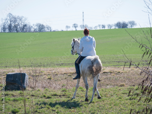 Young sport woman, redhead girl riding on white horse walking backwards at green spring field at city. Seen from back © Kristyna