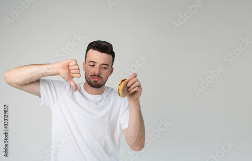 Young man isolated over background. Guy hold big thumb down for untasty burger in his hand. Cheeseburger unhealthy and fat. Bearded man in white shirt stand alone in studio. photo