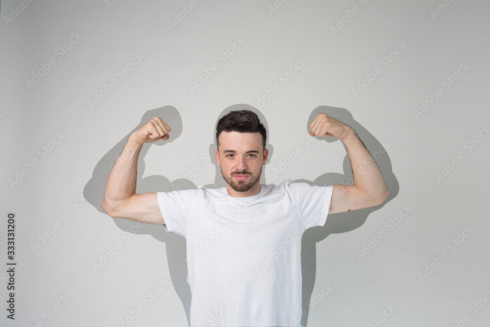 Young man isolated over background. Strong powerful guy show muscled arms. Powerful shadow. Pretend to be strong. Posing on camera alone in studio. Young man isolated over background. Strong powerful guy show muscled arms. Powerful shadow. Pretend to be strong. Posing on camera alone in studio.