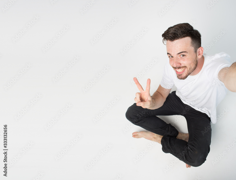 Young man isolated over background. Guy sit on floor and take selfie. Hold camera with one hand and show peace symbol. Emotional happy guy on picture. Young man isolated over background. Guy sit on floor and take selfie. Hold camera with one hand and show peace symbol. Emotional happy guy on picture.