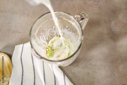 Skim milk is poured into a blender, which contains kiwi, oats and a banana. For a healthy and nutritious smoothie. Concrete background and white striped kitchen towel.