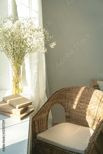 Cozy reading nook by the window with rattan wicker chair, delicate white gypsophila flowers, opened book and window light and shadows.Modern comfortable living room space