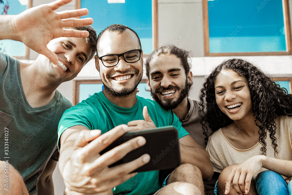 Obraz premium Cheerful friends during video chat via smartphone. Low angle view of happy multiethnic friends sitting together and talking during video chat outdoors. Communication concept