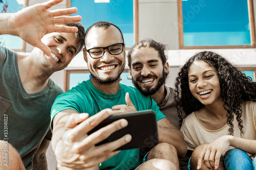 Cheerful friends during video chat via smartphone. Low angle view of happy multiethnic friends sitting together and talking during video chat outdoors. Communication concept © Mangostar