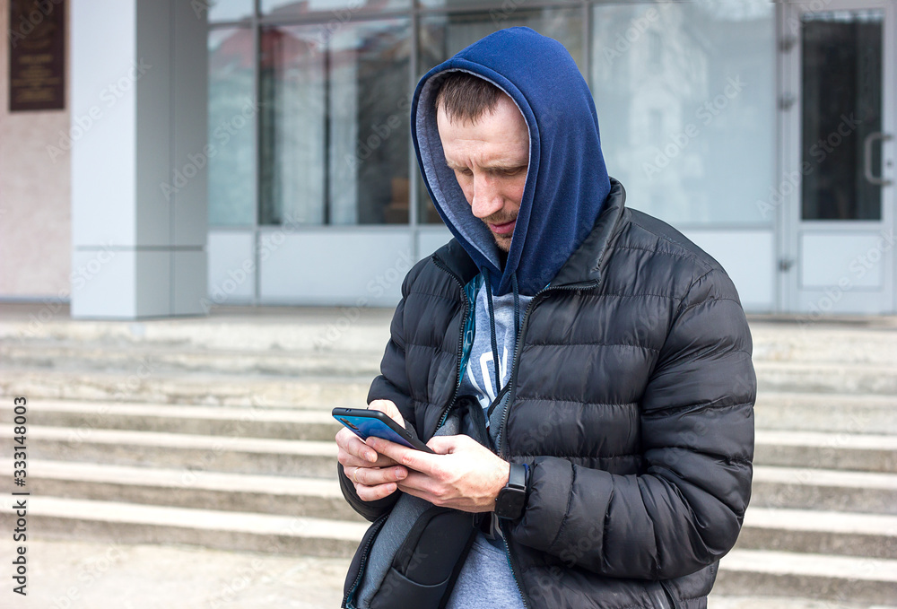 A young man in sportswear and a hood on a city street looks at the phone screen with a smart watch on his hand. The concept of tourism technology and modern lifestyle A young man in sportswear and a hood on a city street looks at the phone screen with a smart watch on his hand. The concept of tourism technology and modern lifestyle