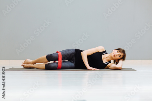 Fototapeta Naklejka Na Ścianę i Meble -  Determined to win. Modern young woman in sport clothing crouching using resistance band while exercising in the gym