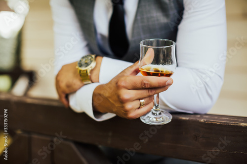 Groom in stylish suits drinking whiskey in hotel room, morning before the wedding preparation, emotional morning. Close up of a man sitting at a table having a glass of cognac. 