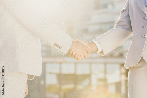 Business colleagues greeting each other in early morning. Business man and woman shaking hands in sunlight. City buildings in background. Agreement concept