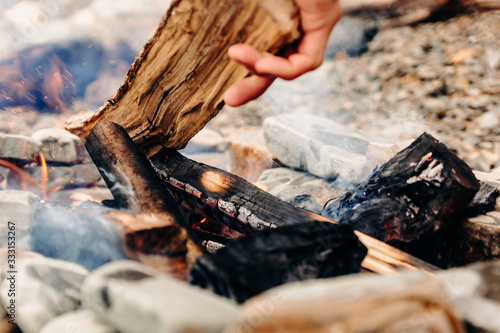 A man throws firewood into a bonfire burning on a stone beach