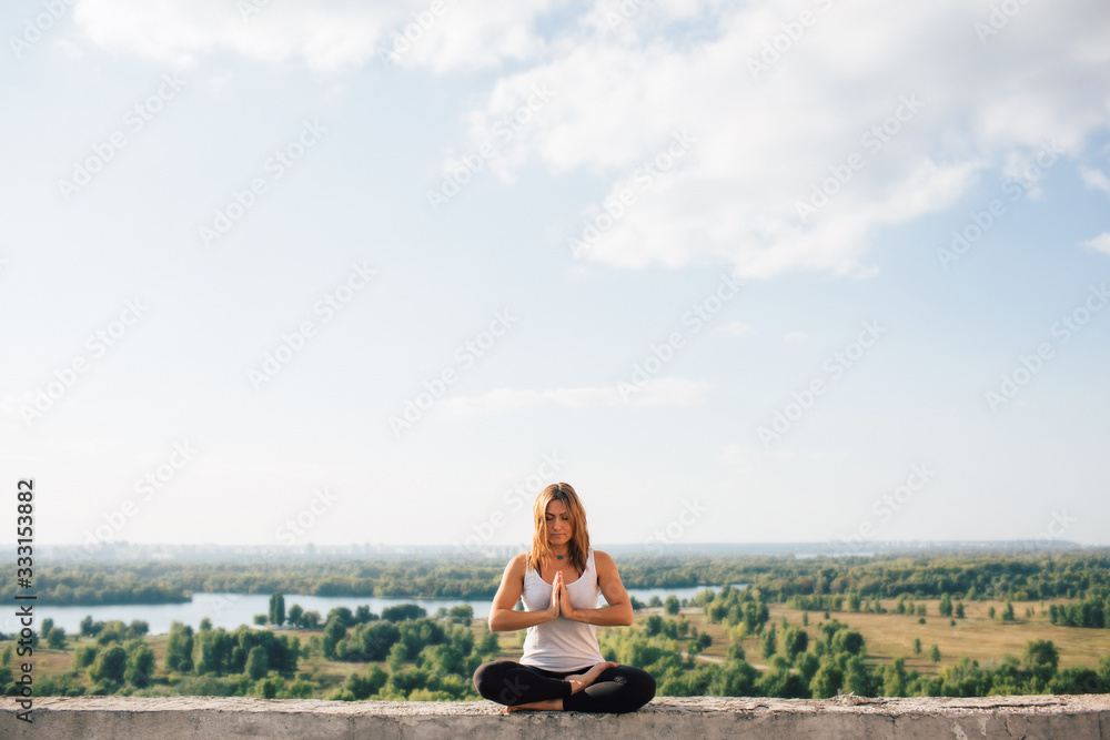 Young woman practices yoga outside. Calm peaceful girl sitting on parapet in lotus position. Her hands at chest level touch each other. Trees river and sky on background.
