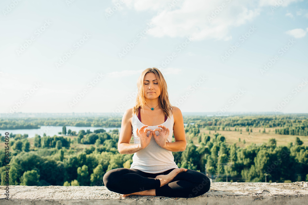 Young woman practices yoga outside. Calm smiling girl sits on parapet in lotus position with close eyes. Her fingers fold into sign of infinity. Nature trees river and sky on background.