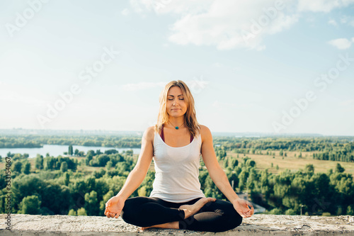 Young woman practices yoga outside. Calm smiling girl sitting on parapet in lotus position with closed eyes. Her hands lowered to knees. Trees river and sky on background.