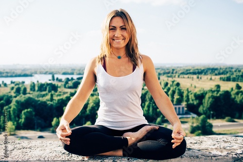 Young woman practices yoga outside. Calm smiling girl sitting on parapet in lotus position with open eyes. Her hands lowered to knees. Trees river and sky on background.