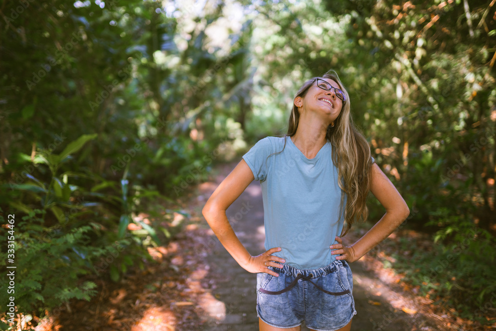 Fototapeta premium A beautiful and emotional girl rejoices among the tropical and exotic forest or jungle on the island of Bali. She shows her different emotions with tanned skin and glasses.