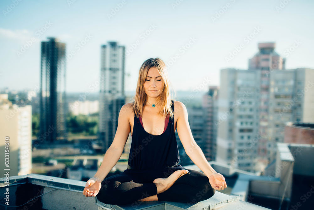 Young woman practices yoga outside. Calm smiling girl sitting on rooftop in lotus position with closed eyes. Her hands lowered to knees. City and high-rise buildings on background. Young woman practices yoga outside. Calm smiling girl sitting on rooftop in lotus position with closed eyes. Her hands lowered to knees. City and high-rise buildings on background.