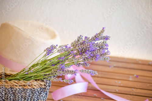Light straw hat on wooden brown table. Lilac-pink ribbon on hat of woman's hat. A bouquet of dry lavender in rustic wicker straw basket.