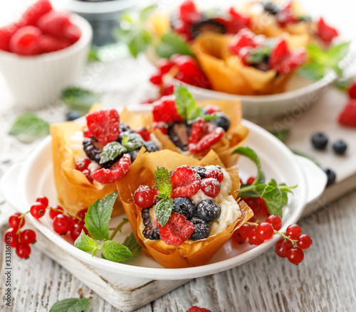 Phyllo pastry fruit cups with whipped cream filling topped with mix fresh berries sprinkled with powder sugar on a white plate close up. 