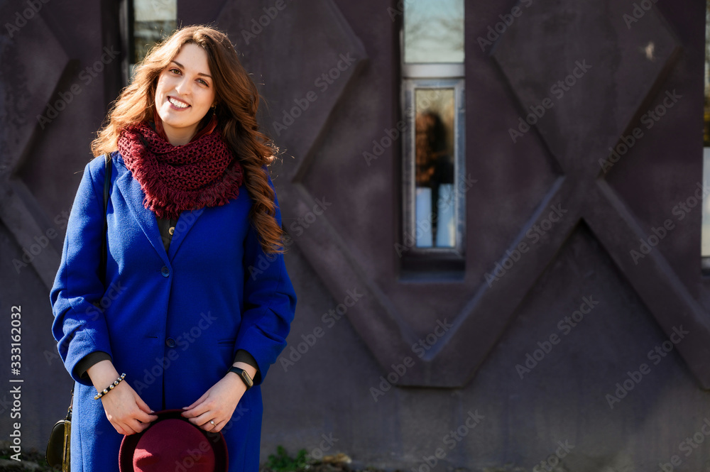 Portrait of a pretty fashionable brunette woman with long beautiful hair in a blue coat on a background of a building wall
