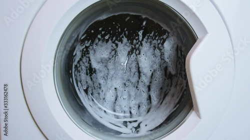 Washing clothing in generic domestic washing machine.Close up video of spinning washing machine drum with foam spraying door. 