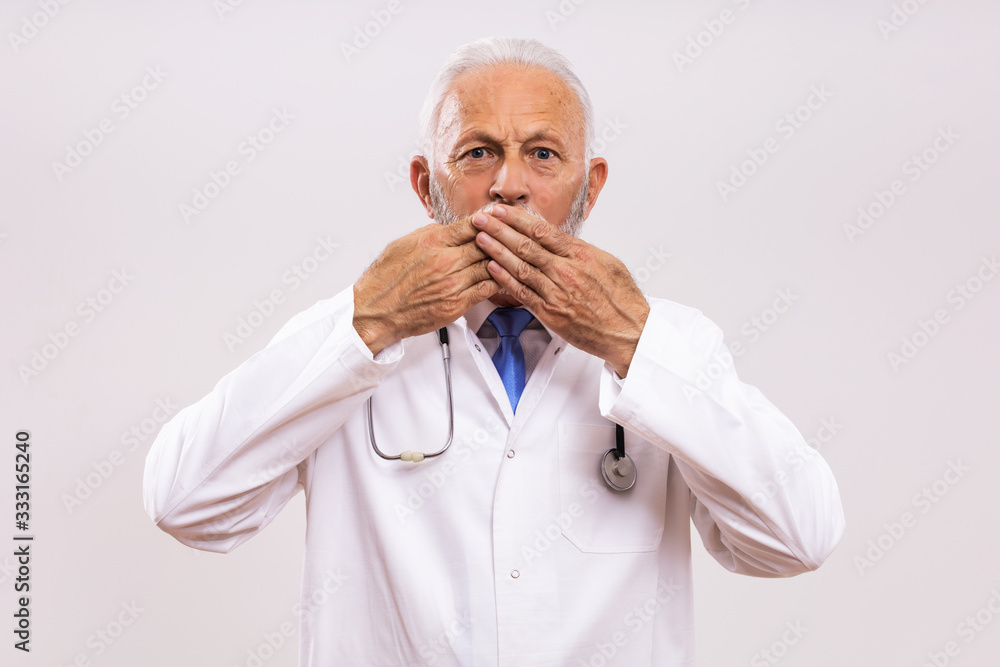 Shocked senior doctor with hands covering mouth on gray background. Shocked senior doctor with hands covering mouth on gray background.