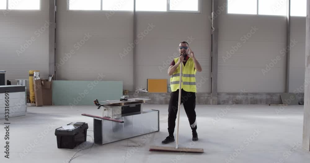 A cheerful and well-disposed worker dances with the sweeper while cleaning the deposit. Relaxation while working, fun on the job site listening to music. A man who loves his profession. A cheerful and well-disposed worker dances with the sweeper while cleaning the deposit. Relaxation while working, fun on the job site listening to music. A man who loves his profession.