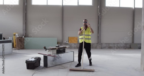 A cheerful and well-disposed worker dances with the sweeper while cleaning the deposit. Relaxation while working, fun on the job site listening to music. A man who loves his profession.
