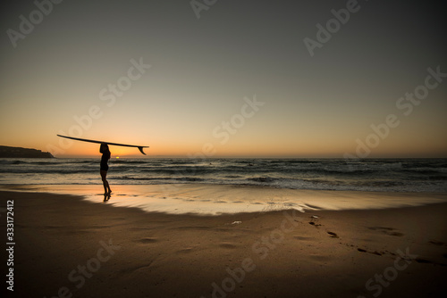 Female surfer standing on beach at sunrise holding a surfboard