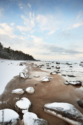 A view of the snow-covered Baltic sea coast at sunset. Stones in the water close-up. Coniferous forest in the background. Stunning cloudscape. Warm evening light. Kaltene, Latvia photo