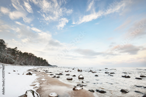 A view of the snow-covered Baltic sea coast at sunset. Stones in the water close-up. Coniferous forest in the background. Stunning cloudscape. Warm evening light. Kaltene, Latvia photo